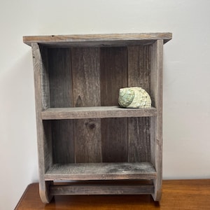 May include: A rustic, wooden shelf with two shelves and a towel bar. The shelf is made of weathered, gray-brown wood. A decorative seashell rests on the top shelf. The shelf is designed for wall mounting and adds a touch of coastal decor.