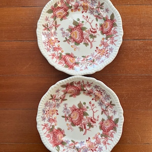 May include: Two white ceramic plates with a floral design. The plates feature a scalloped edge and a pattern of red, pink, and yellow flowers with green leaves. The plates are on a wooden surface.