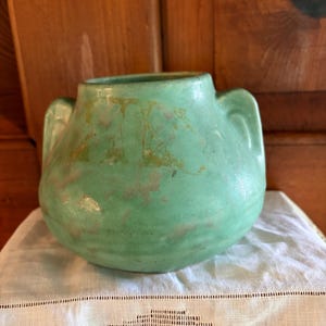 May include: A round, light green ceramic vase with two side handles. The surface features a matte finish with subtle, mottled brown and tan discoloration across the body. The vase has a wide, open mouth and a rounded base, showing signs of age.