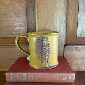 May include: A yellow ceramic mug with a brown handle and rim, featuring embossed text. The mug is placed on top of two red books. The text on the mug reads: "ENGENCERING, TELLERICA MASS, APRIL 22, 1969".