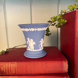May include: A light blue ceramic vase with white relief figures and a decorative rim. The vase is set on a stack of red books. The books have gold lettering and the text "The American Claimant" and "Tom Sawyer Abroad" are visible.