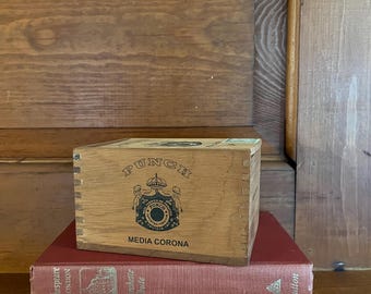 May include: A wooden box with the words "PUNCH MEDIA CORONA" printed on the front. The box is sitting on top of a stack of three books.