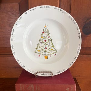 May include: A white ceramic plate featuring a central illustration of a decorated Christmas tree with a gold star topper. The rim is inscribed with the repeating text all is Calm and all is Bright in a black, handwritten-style font.
