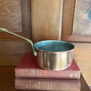 May include: A copper saucepan with a brass handle sits atop two antique books with red covers. The pan has a shiny exterior and a green interior. The books are titled "Shakespeare London" and "The Days of Henry Thoreau."