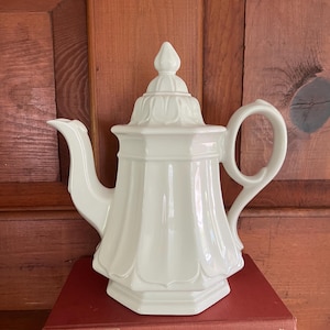 May include: A white ceramic teapot with a lid, spout, and handle. The teapot has a fluted design and sits on top of a stack of red books. The background is a wooden door.