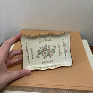 May include: A rectangular cream-colored ceramic trinket tray with gold-trimmed scalloped edges. The center features a floral motif and the text: Love is Patient, never proud, never boastful, Love is Kind. The tray has decorative floral borders.