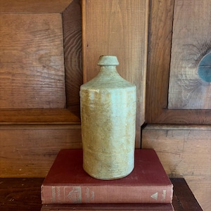 May include: A cylindrical stoneware bottle with a light beige, mottled glaze and a narrow neck featuring a prominent rim. The bottle has a smooth, slightly uneven texture and a sturdy base, standing upright with a simple, functional design.