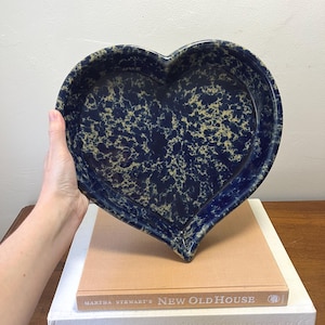 May include: A navy blue heart-shaped ceramic serving dish with a speckled pattern in tan and blue. The dish is resting on top of two books, one titled "Martha Stewart's New Old House."