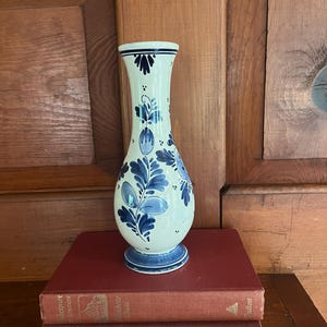 May include: A white and blue ceramic vase with a floral design. The vase has a flared top and a round base. The floral design is in shades of blue. The vase is sitting on top of two red books.