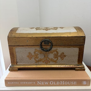 May include: A rectangular wooden chest with a domed lid, featuring a distressed white and gold finish with ornate gold scrollwork patterns. The chest has a dark metal ring pull handle on the front and sits atop a tan book titled New Old House.