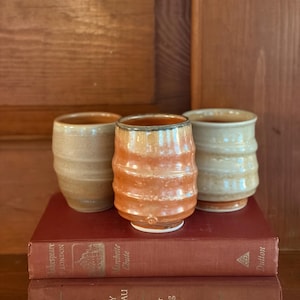 May include: Three ceramic tumblers with a ribbed texture. The center tumbler features a glossy orange glaze, while the two flanking tumblers have a matte beige finish. These vessels are positioned on top of two stacked hardcover books.