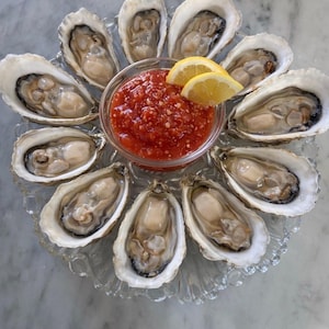 May include: A dozen fresh oysters arranged in a circle on a clear glass plate. A small glass bowl in the center holds a red sauce, garnished with two lemon wedges. The oysters are open, revealing the oyster meat.