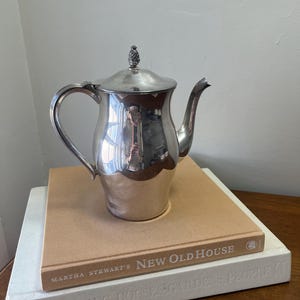 May include: A silver teapot with a lid and a curved spout, sitting on top of two books. The teapot has a reflective surface and a decorative finial on the lid. The books are titled "New Old House" and "Vogue Living Houses Gardens People".