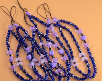 May include: Multiple beaded phone charms in a deep blue hue, decorated with lavender heart and butterfly charms. They have black cords for attachment and are set against a light orange backdrop.