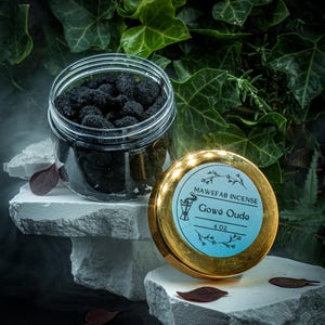 May include: A clear jar filled with dark incense pieces sits next to a gold-lidded container. The lid has a blue label with the text "MAWEFAB INCENSE Gowé Oude 4 oz". The background features green foliage and white stone platforms.