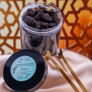 May include: A clear plastic jar filled with dark brown incense, a black lid with a label that reads "MAWEFAB INCENSE Gowé 10 OZ", and gold-coloured tongs with heart-shaped ends. The items are on a peach-coloured fabric.