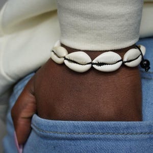 May include: A bracelet made of white cowrie shells strung together with black string. The bracelet is worn on a dark-skinned wrist, with a white sweater sleeve visible above. The person is wearing blue jeans.