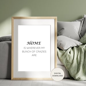 May include: A framed print with the text "HOME IS WHEREVER MY BUNCH OF CRAZIES ARE" in black text on a white background. The frame is light wood and the print is sitting on a light green blanket.