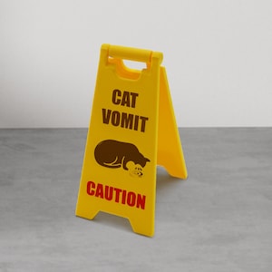 以下が含まれることがあります： 「CAT VOMIT」と茶色で、「CAUTION」と赤色で書かれた、明るい黄色の折りたたみ式注意看板。茶色の猫のグラフィックが、少量の嘔吐物とともに表示されています。この看板は自立するように設計されています。