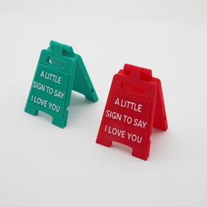 Puede incluir: Dos pequeños letreros en forma de A, uno verde y otro rojo, cada uno con el mensaje "A LITTLE SIGN TO SAY I LOVE YOU" en texto blanco. Los letreros están hechos de un material sólido y se mantienen en pie.