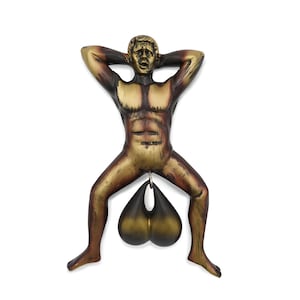 May include: A bronze-colored sculpture of a nude male figure with arms behind his head and legs spread. A dark, teardrop-shaped object hangs from the figure's groin. The sculpture has a distressed finish and is set against a white background.