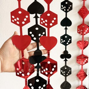 May include: Red and black playing card-themed garland. The garland features hearts, spades, clubs, and dice shapes. The dice have white dots. The garland is made of a shiny, reflective material.
