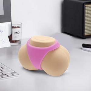 Puede incluir: Un accesorio de escritorio liso de color beige con forma de glúteos que llevan una tanga de color rosa brillante. El objeto tiene una forma redondeada y ergonómica con una superficie superior plana, diseñado para colocarse sobre un escritorio.