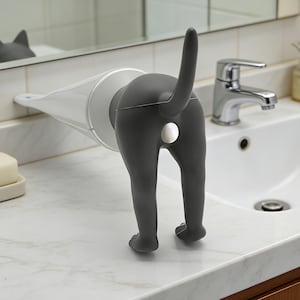 May include: A novelty cat-shaped faucet extender in gray, attached to a white and gray conical water spout. The cat's rear end and tail are prominently displayed, with a silver-colored circle. The item is mounted on a white marble countertop.