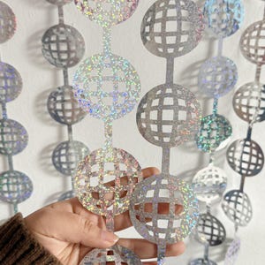 May include: A string of silver disco ball decorations with a holographic, glittery finish. The cut-out spheres are connected by thin, vertical strips. The decorations are ideal for parties and celebrations.