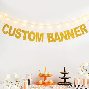 May include: A gold glitter "CUSTOM BANNER" strung across a white wall, illuminated by a string of lights. The banner is above a table set for a party, with cupcakes, plates, and balloons.