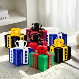 Puede incluir: Una colección de seis recipientes cuadrados con forma de caja de regalo y lazos en la parte superior. Las cajas vienen en varias combinaciones de colores, incluyendo negro con lazos amarillos, rojo con lazos negros, azul con lazos blancos y verde con lazos rojos.