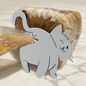Puede incluir: Un clip para pan con forma de gato gris, diseñado para sellar bolsas de pan. El clip está hecho de un material gris claro sólido y presenta un diseño de gato caprichoso con cola y cara sonriente. El clip sujeta una bolsa de pan de plástico transparente.