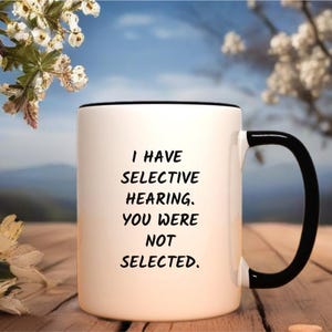May include: White ceramic coffee mug with a black rim and handle. The mug has black text that reads "I have selective hearing. You were not selected."