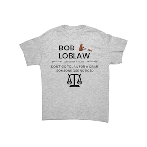 May include: A light gray t-shirt with the text "BOB LOBLAW ATTORNEY AT LAW" and the phrase "DON'T GO TO JAIL FOR A CRIME SOMEONE ELSE NOTICED". The design includes a gavel and scales.