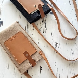 Puede incluir: Dos fundas para teléfono de fieltro con detalles de cuero y correas de cuero ajustables. Las fundas son beige y negra y ambas tienen la palabra "Ecolution" grabada en el cuero.