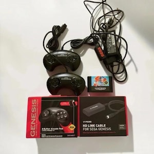 May include: A Sega Genesis gaming bundle including two black controllers, a game cartridge, a power adapter, and a Pound HD Link cable. The set features two retail boxes, one for a 6-button arcade pad and one for the HD link cable.