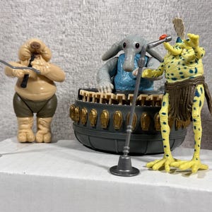 Può includere: Tre action figure di musicisti alieni. Una figura è color camoscio con pantaloncini marroni, un'altra è grigia con un gilet blu e la terza è gialla con macchie blu e una gonna marrone. Ogni figura ha un microfono davanti.