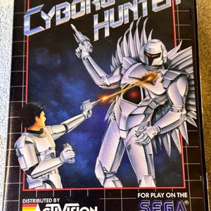 May include: A vintage SEGA Master System game box for "Cyborg Hunter." The cover art features two cyborg figures in silver armor, one firing a weapon. The background is a dark blue with the Activision logo.