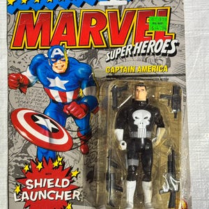 Könnte beinhalten: Marvel Super Heroes Captain America und The Punisher Actionfiguren in Originalverpackung. Die Verpackung zeigt Captain America mit seinem Schild und dem Text "SHIELD LAUNCHER". Die Punisher-Figur ist schwarz-weiß mit mehreren Waffen.