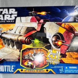 May include: A Star Wars: The Clone Wars Republic Attack Shuttle toy box. The box features a detailed image of the shuttle with electronic lights, sounds, and phrases. It includes a Clone Pilot figure and converts to an attack base.