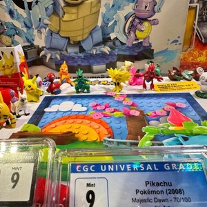 Puede incluir: Una exhibici&oacute;n de varias figuras de Pok&eacute;mon, incluido Pikachu, y juegos de construcci&oacute;n. La escena incluye cartas calificadas con una calificaci&oacute;n "Mint 9" y un colorido tel&oacute;n de fondo de paisaje.