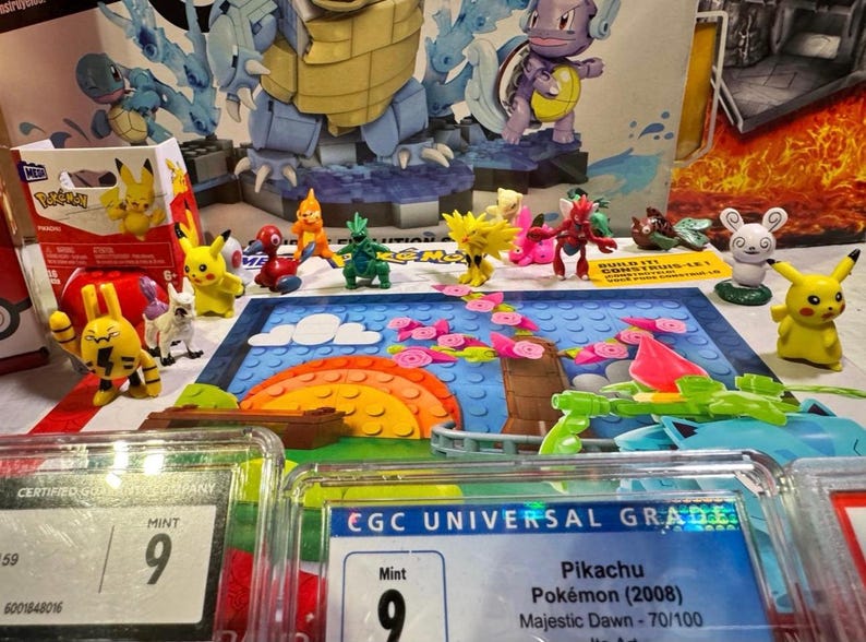 Puede incluir: Una exhibici&oacute;n de varias figuras de Pok&eacute;mon, incluido Pikachu, y juegos de construcci&oacute;n. La escena incluye cartas calificadas con una calificaci&oacute;n "Mint 9" y un colorido tel&oacute;n de fondo de paisaje.