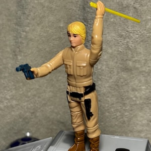 May include: A vintage Star Wars action figure, featuring a character in a tan uniform and brown boots. The figure holds a blue blaster and a yellow lightsaber. It stands on a grey surface. A classic collectible toy.