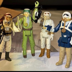 Puede incluir: Cuatro figuras de acción vintage de una franquicia de ciencia ficción. Las figuras son de varios colores, incluyendo blanco, verde y azul, y llevan cascos y equipo. Cada figura sostiene un arma o accesorio, como una ballesta o un bláster.