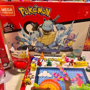 Puede incluir: Un juego de construcci&oacute;n Mega Construx Pok&eacute;mon rojo y negro con una gran figura de Blastoise con efectos de agua. La caja muestra el logotipo de Pok&eacute;mon y varias figuras m&aacute;s peque&ntilde;as, incluyendo Pikachu y Squirtle.
