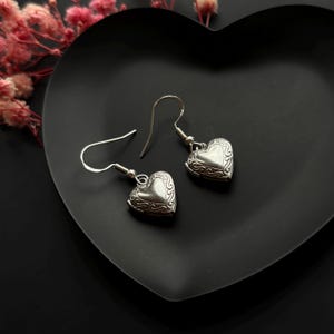 Puede incluir: Un par de pendientes de medallón en forma de corazón de tono plateado con intrincados patrones de volutas en relieve en los bordes. Cada medallón cuelga de un sencillo gancho de metal plateado para orejas perforadas, con acabado pulido.