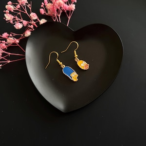 May include: A pair of gold-toned hook earrings featuring enamel charms of two cartoon characters. One charm depicts a figure with tall blue hair, while the other charm shows a yellow-skinned character with a prominent mouth and wide eyes.
