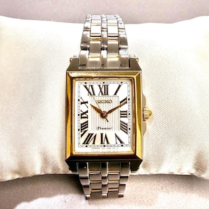 May include: A rectangular Seiko Premier watch with a gold-tone bezel and a silver-tone band. The white watch face features black Roman numerals and gold hands. The watch is displayed on a white cushion.