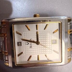 May include: A rectangular Bulova wristwatch with a two-tone silver and gold metal band. The watch face features a silver grid pattern, gold-tone hour markers, gold hands, a date window at the three o'clock position, and a single diamond accent.