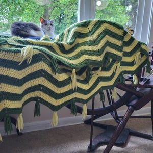 May include: A handmade crochet blanket with a chevron pattern in green and yellow. The blanket has tassels along the bottom edge. A cat is resting on a window sill in the background.